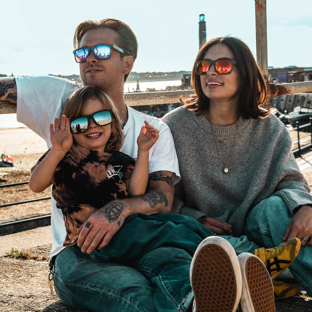 a family of three sit beside the beach on a sunny day wearing matching sunglasses